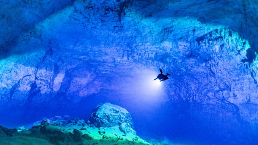 Massive underwater cave chamber in Malazamanga Cave, Madagascar, illuminated by three cave divers with powerful dive lights.